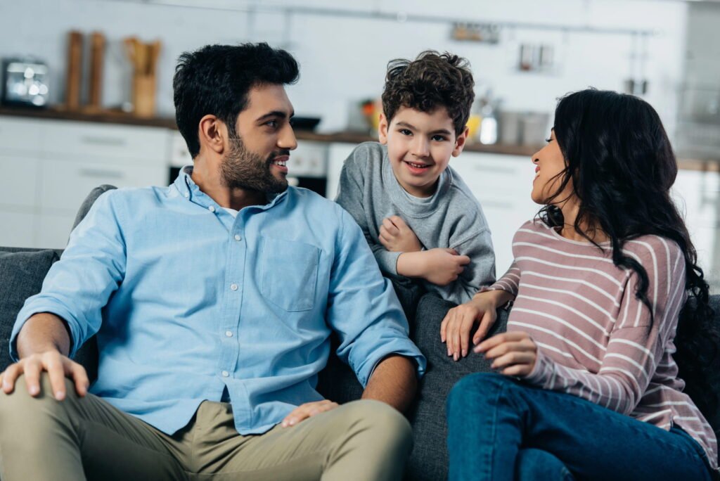 Parents talking on a couch while their son comes in between them from behind the couch